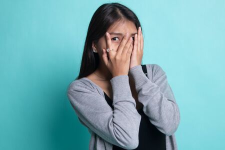 Beautiful young Asian woman peeking through fingers on cyan background.の写真素材