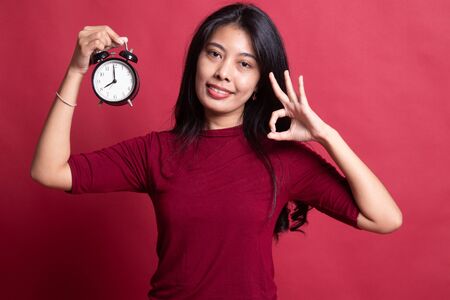 Young Asian woman show OK with a clock on red background.