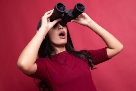 Young Asian woman with binoculars on red background.