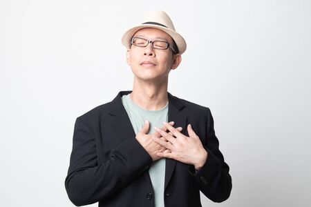 Faithful  adult asian man closes eyes and keeps hands on chest near heart, shows her kindness on white backgroundの写真素材