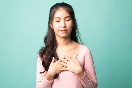 Faithful woman closes eyes and keeps hands on chest near heart, shows her kindness on cyan backgroundの写真素材