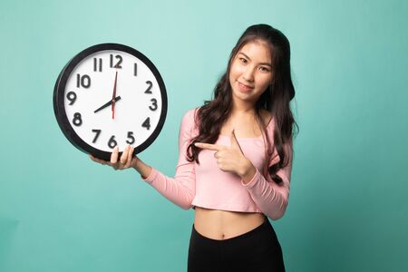 Young Asian woman point to a clock on cyan backgroundの写真素材