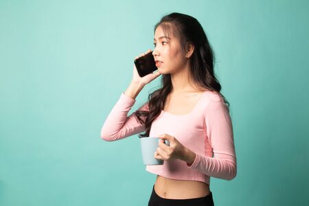 Young Asian woman with mobile phone and coffee cup on cyan backgroundの写真素材