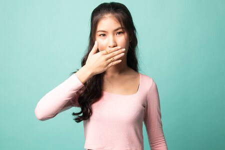 Young Asian woman close her mouth with hand on cyan backgroundの写真素材