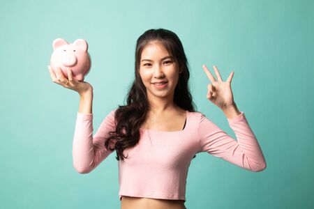Asian woman show OK  with pig coin bank on cyan backgroundの写真素材