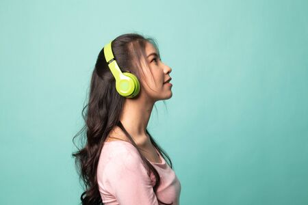 Pretty asian girl listening music with her headphones on cyan backgroundの写真素材