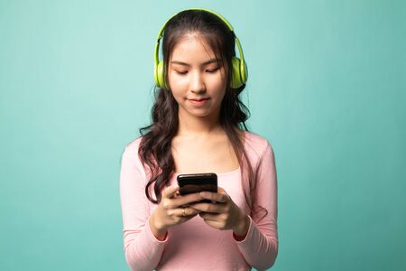 Pretty asian girl listening music with her headphones on cyan backgroundの写真素材