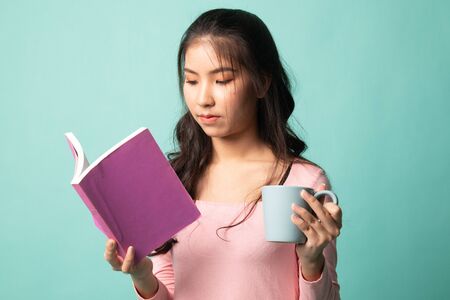 Young Asian woman with a book and cup of coffee on cyan backgroundの写真素材