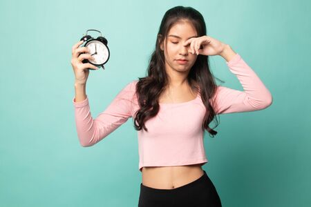 Sleepy young Asian woman with a clock in the morning on cyan backgroundの写真素材
