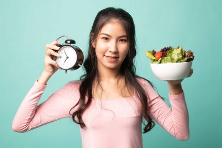 Young Asian woman with clock and salad on cyan backgroundの写真素材
