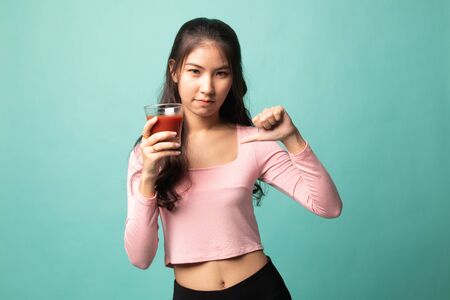 Young Asian woman  thumbs down hate tomato juice on cyan backgroundの写真素材