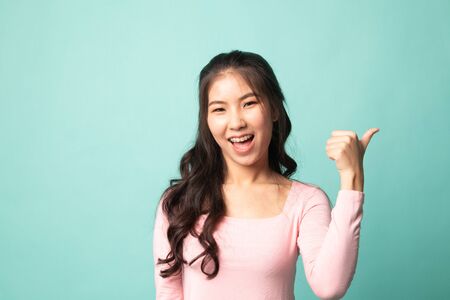 Young Asian woman smile and point to copy space with her thumb on cyan backgroundの写真素材