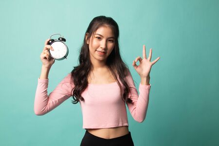 Young Asian woman show OK with a clock on cyan backgroundの写真素材