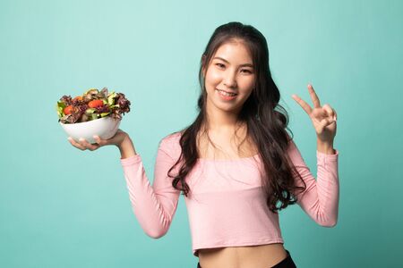 Healthy Asian woman show victory sign with salad on cyan backgroundの写真素材