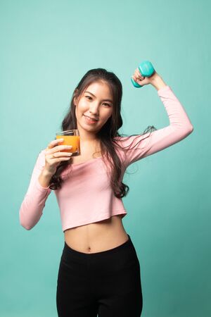 Young Asian woman with dumbbell drink orange juice on cyan backgroundの写真素材