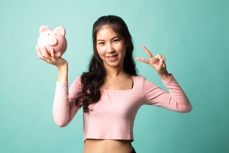 Asian woman show victory sign with pig coin bank on cyan backgroundの写真素材