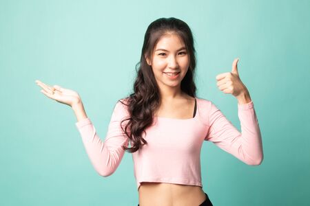 Beautiful young Asian woman show palm hand and thumbs up on cyan backgroundの写真素材