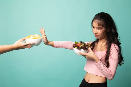 Young Asian woman with salad say no to potato chips on cyan backgroundの写真素材