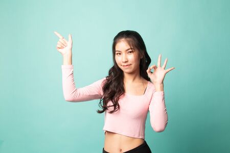 Young Asian woman point and show OK on cyan backgroundの写真素材