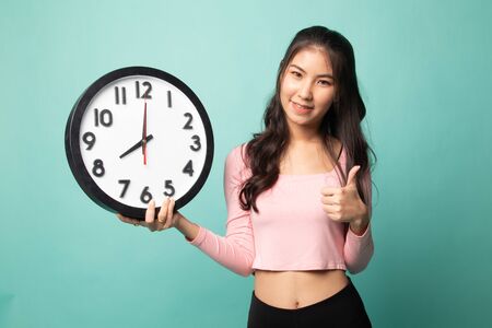 Young Asian woman thumbs up with a clock on cyan backgroundの写真素材