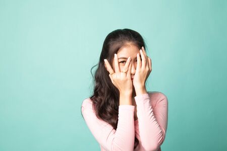 Beautiful young Asian woman peeking through fingers on cyan backgroundの写真素材