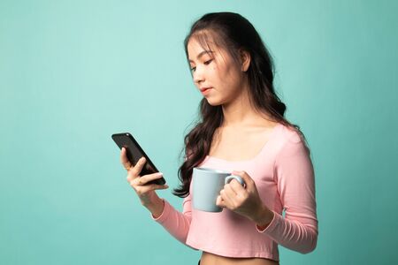 Young Asian woman with mobile phone and coffee cup on cyan backgroundの写真素材