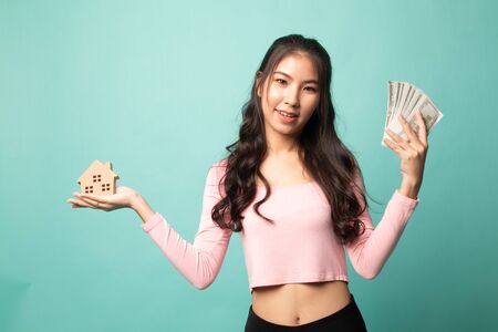Happy young asian woman with house model and bunch of money banknotes on cyan backgroundの写真素材