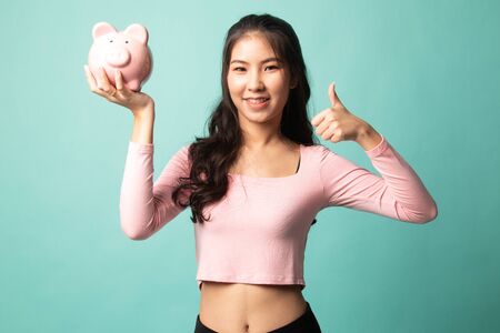 Asian woman thumbs up with pig coin bank on cyan backgroundの写真素材