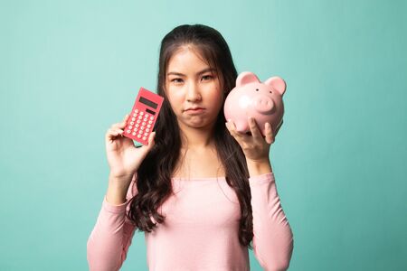 Unhappy Asian woman with calculator and piggy bank on cyan backgroundの写真素材