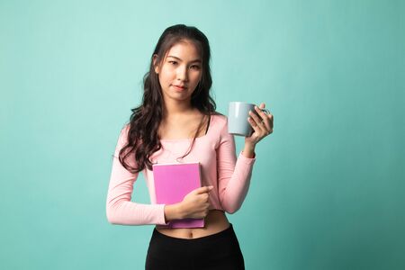 Young Asian woman with a book and cup of coffee on cyan backgroundの写真素材