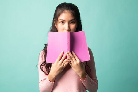Young Asian woman with a book cover her face on cyan backgroundの写真素材