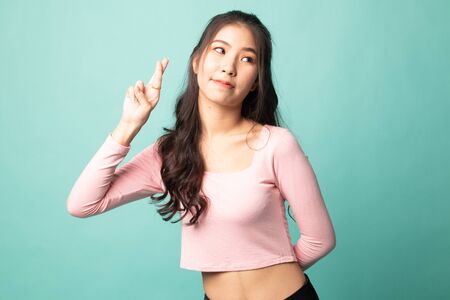 Beautiful young Asian woman cross finger on cyan backgroundの写真素材