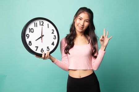 Young Asian woman show OK with a clock on cyan backgroundの写真素材