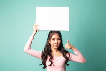 Young Asian woman show thumbs up with white blank sign on cyan backgroundの写真素材
