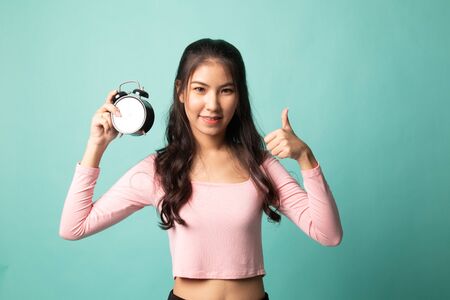 Young Asian woman show thumbs up with a clock on cyan backgroundの写真素材