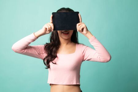 Young Asian woman with a computer tablet over her face on cyan backgroundの写真素材
