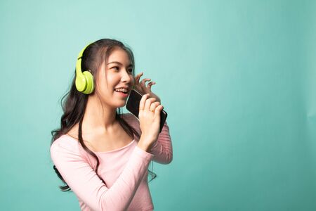 Pretty asian girl listening music with her headphones on cyan backgroundの写真素材