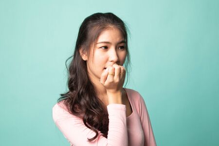 Unsure hesitant nervous young asian  woman biting her fingernails on cyan backgroundの写真素材
