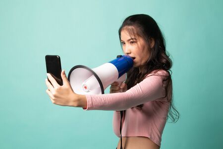 Angry  young Asian woman shout with megaphone to mobile phone on cyan backgroundの写真素材
