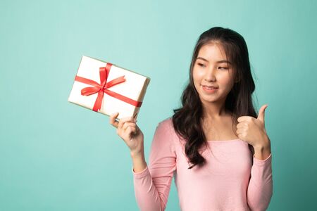 Young Asian woman thumbs up with a gift box on cyan backgroundの写真素材