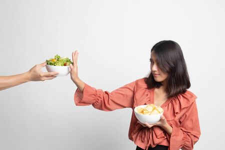 Young Asian woman with potato chips say no to salad on white backgroundの写真素材