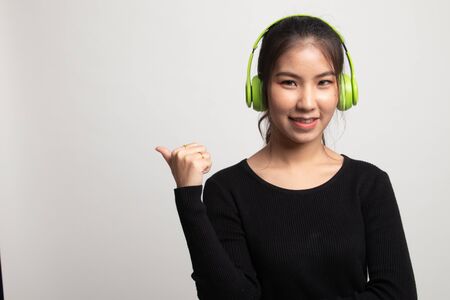 Pretty asian girl listening music with her headphones point to copy space with her thumb on white backgroundの写真素材
