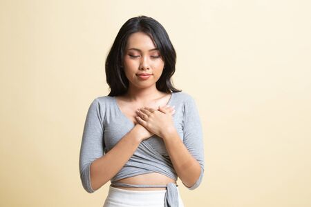 Faithful woman closes eyes and keeps hands on chest near heart, shows her kindness   on beige backgroundの写真素材