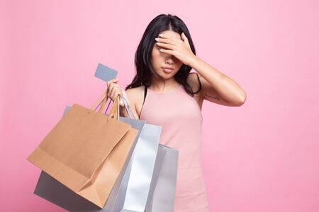 Unhappy  young Asian woman with shopping bags and credit card on pink backgroundの写真素材