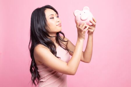 Young Asian woman kiss  a pink coin bank on pink backgroundの写真素材