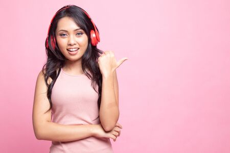Pretty asian girl listening music with her headphones point to copy space with her thumb on pink backgroundの写真素材