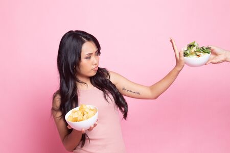Young Asian woman with potato chips say no to salad on pink backgroundの写真素材