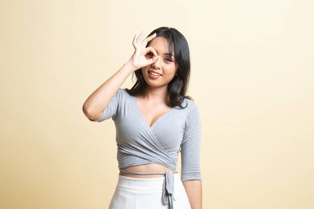 Beautiful young Asian woman show OK sign over her eye   on beige backgroundの写真素材