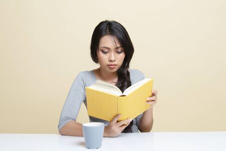 Young Asian woman read a book with cup of coffee   on beige backgroundの写真素材