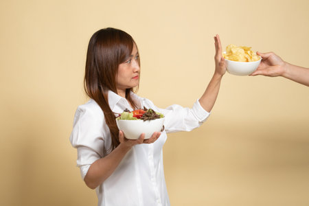 Young Asian woman with salad say no to potato chips on beige backgroundの写真素材